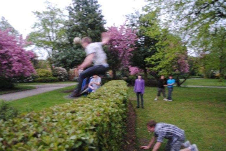 A group stands in a park as a person jumps over a hedge